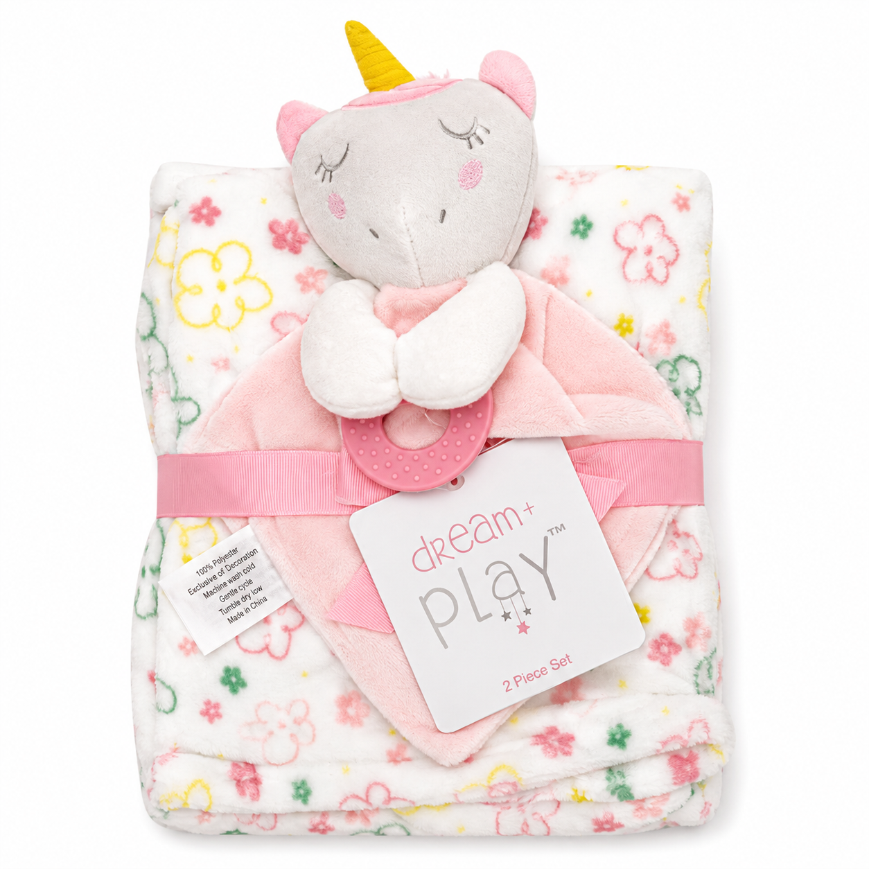A baby comforter set featuring a folded white blanket with pink, yellow, and green floral patterns, a plush unicorn head topper with a yellow horn, a pink teething ring, and a pink ribbon with a 'dream play' tag.
