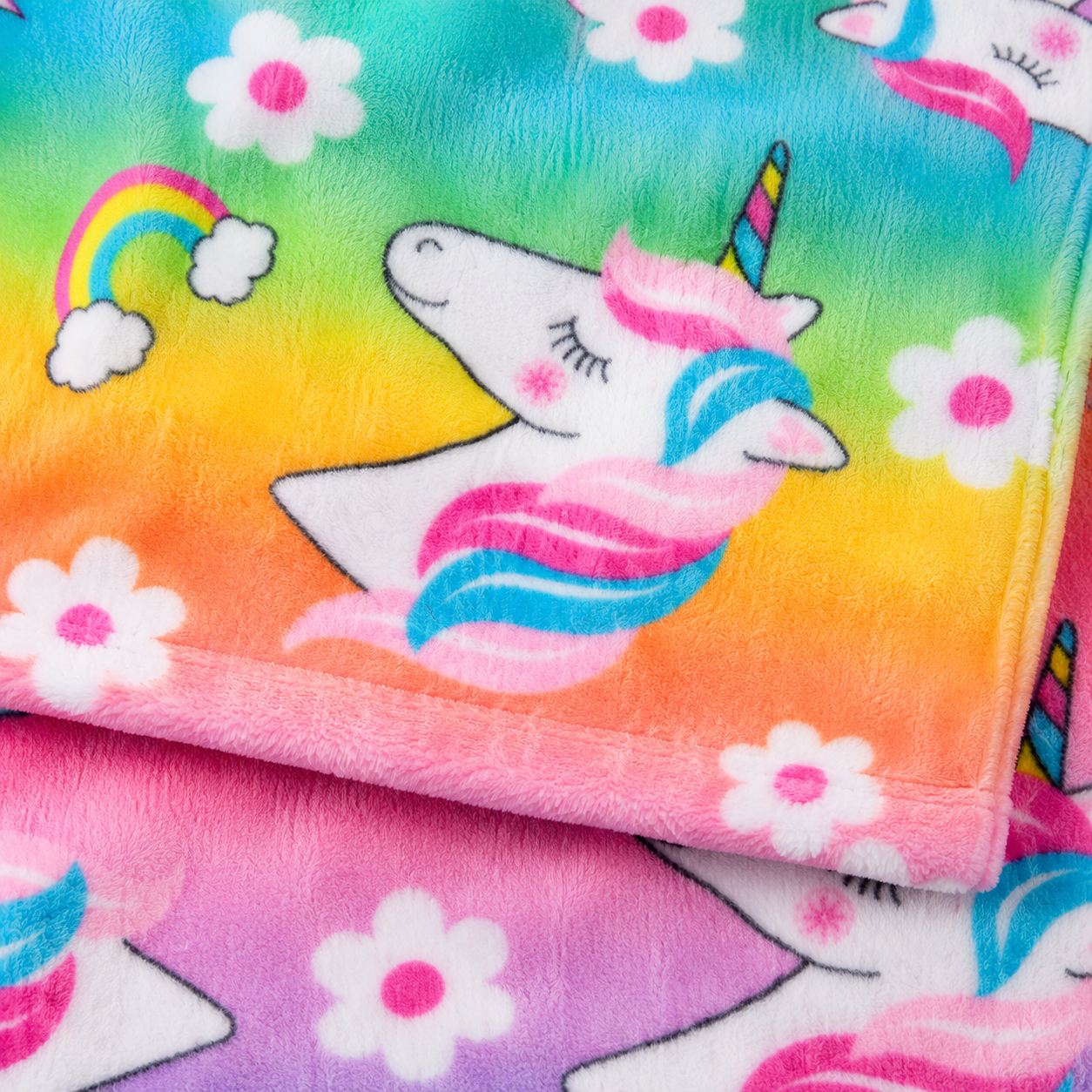 A close-up of a plush blanket with a colorful rainbow gradient background, featuring a white unicorn with pink and blue hair, white flowers, and a small rainbow.