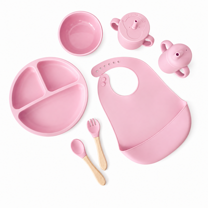 Pink baby feeding set arranged on a white background, including a divided plate, silicone bib, bowl, sippy cup, small cup, spoon, and fork with light wood handles.