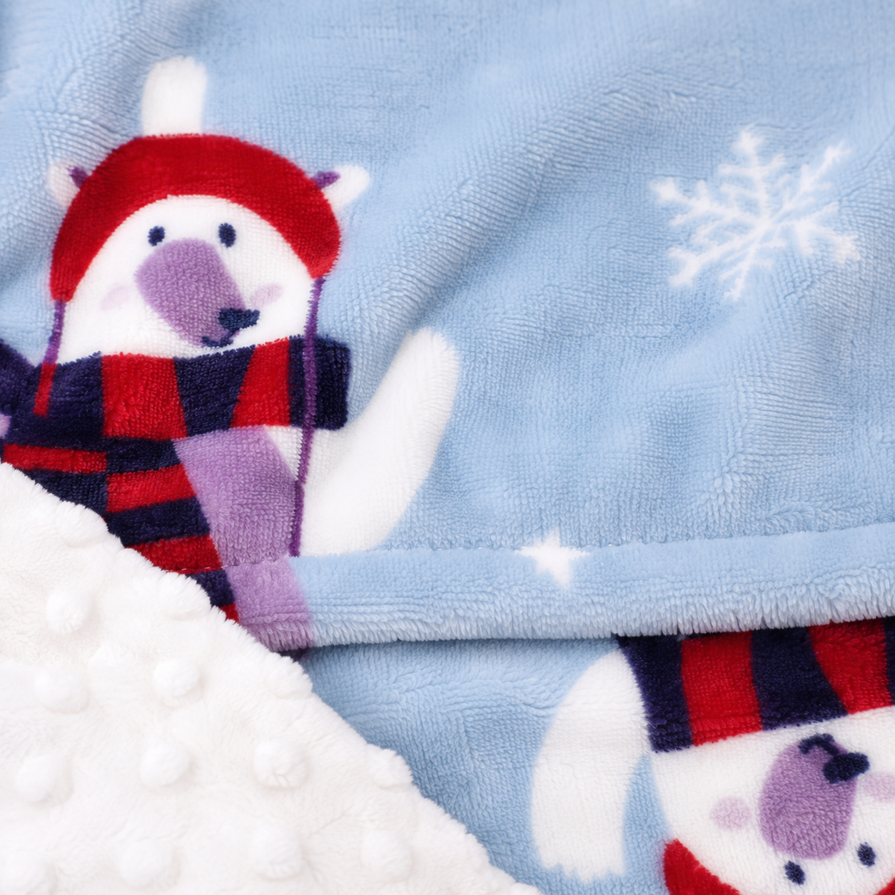 Close-up of a plush blanket with a light blue main fabric, white faux-fur trim, and a cartoon polar bear wearing a red hat and red-and-navy plaid scarf. White snowflake motifs appear on the blue fabric.