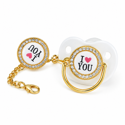 Gold baby pacifier clip with a matching clip-on pacifier. The pacifier features a white shield with the text 'I [heart] YOU' in black with a pink heart, surrounded by a gold bezel set with clear rhinestones.