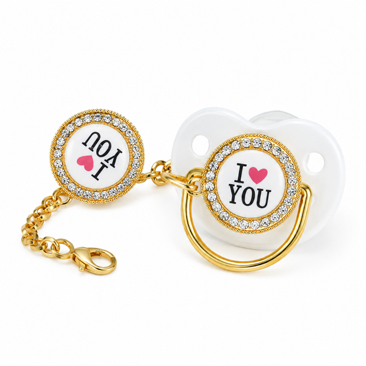 Gold baby pacifier clip with a matching clip-on pacifier. The pacifier features a white shield with the text 'I [heart] YOU' in black with a pink heart, surrounded by a gold bezel set with clear rhinestones.