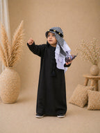 Child in a black outfit with a checkered headscarf standing in a decorated room with plants and pillows.