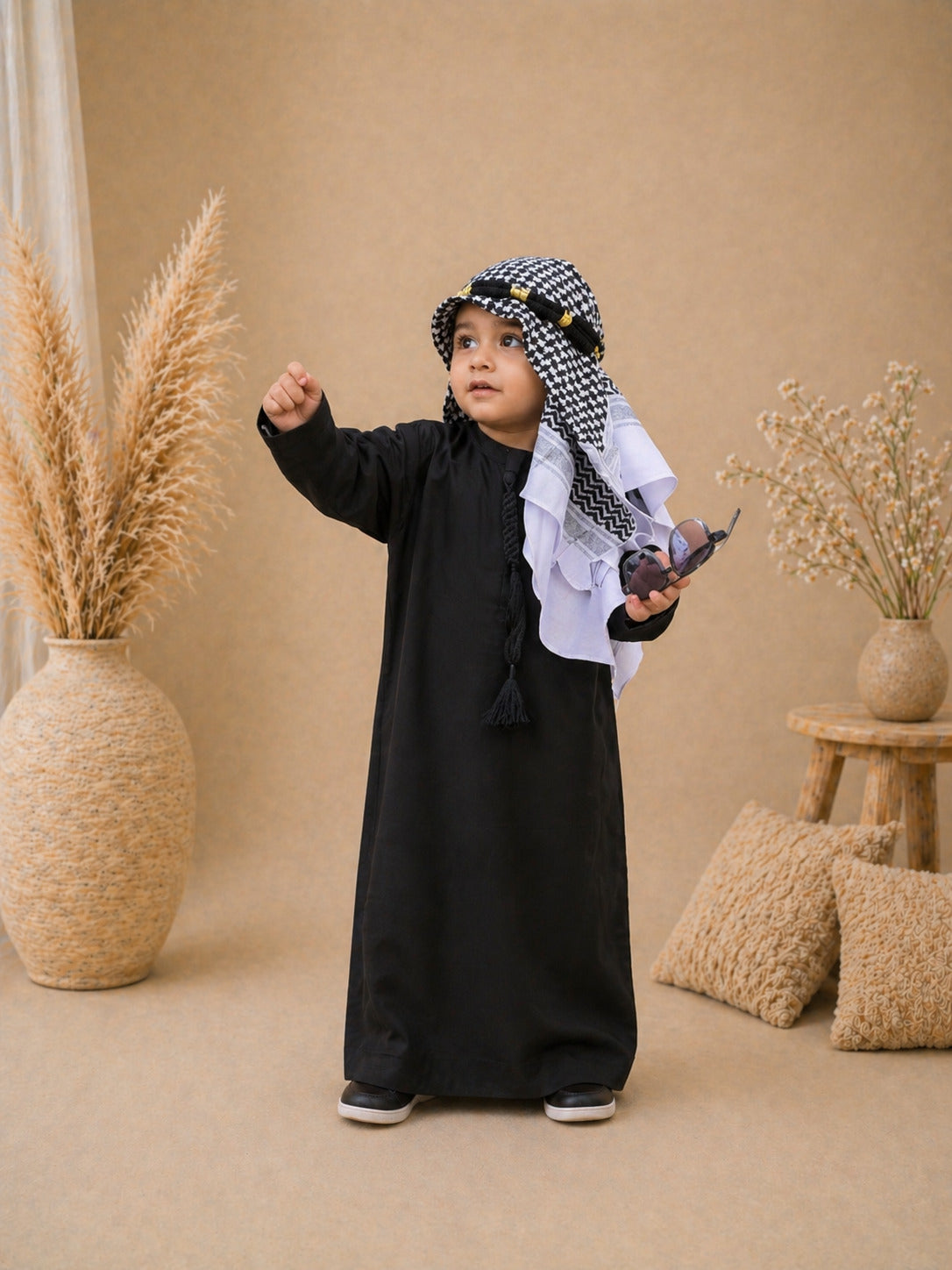 Child in a black outfit with a checkered headscarf standing in a decorated room with plants and pillows.