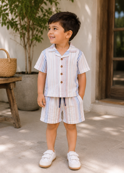 A young boy wearing a matching short-sleeve button-front shirt and shorts set with vertical blue, pink, and white stripes, standing outdoors on a patio.