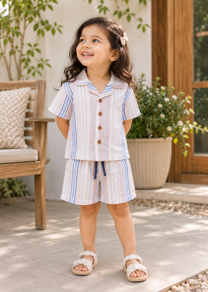 A young girl stands on a patio wearing a matching short-sleeve button-front top and shorts set with vertical light blue and white stripes.