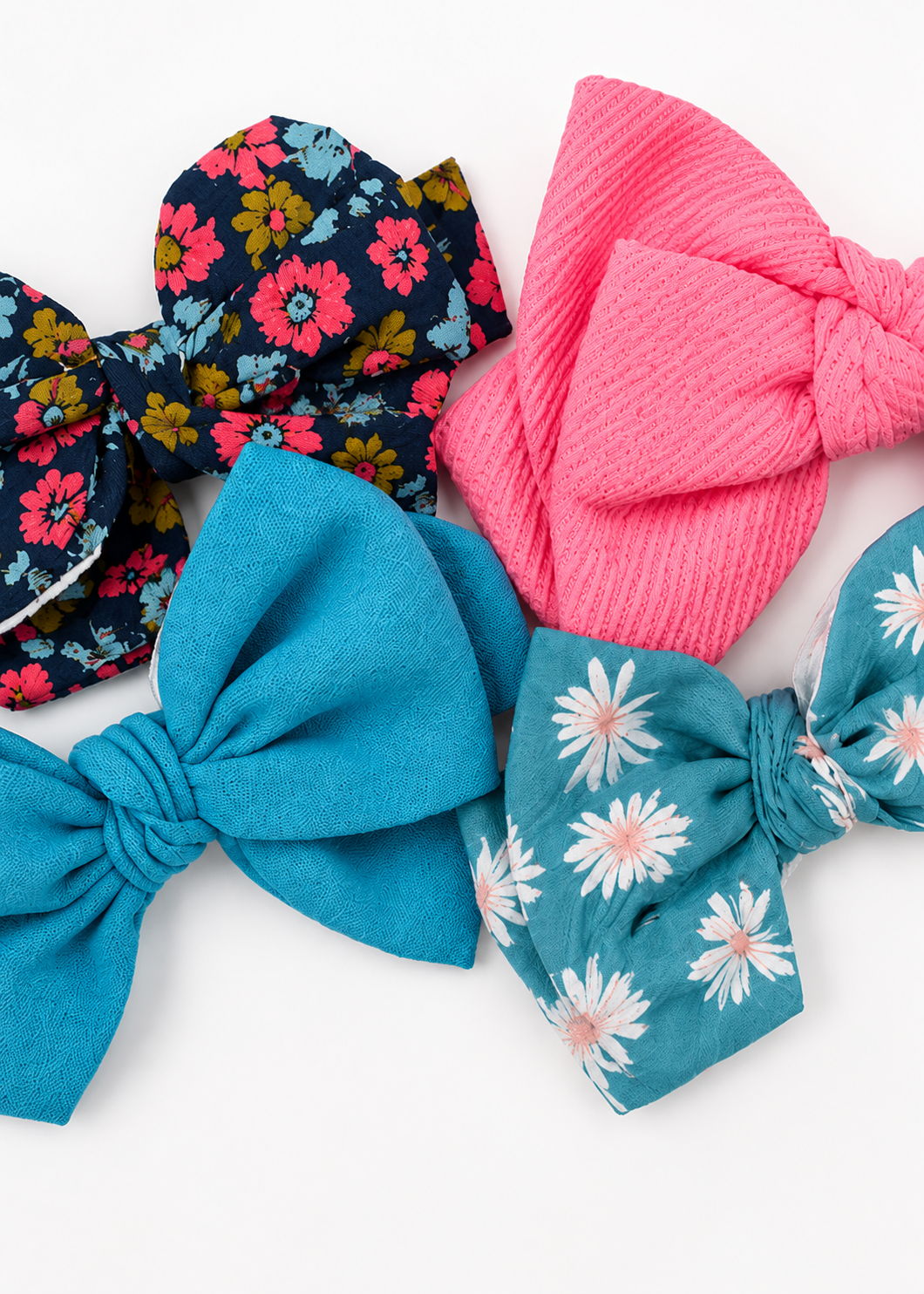 Four fabric hair bows arranged on a white background, including solid bright pink, solid turquoise blue, navy floral with pink and yellow flowers, and teal floral with white daisies.