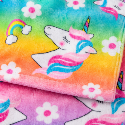 A close-up of a plush blanket with a colorful rainbow gradient background, featuring a white unicorn with pink and blue hair, white flowers, and a small rainbow.